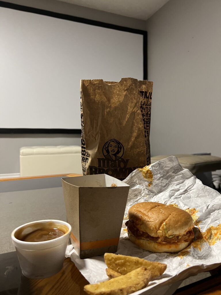 A picture with all the items of a Mary's Buffalo sandwich combo laid out. Pictured from left to right is: gravy in a container, a fry box and some fry's, the Mary's buffalo sandwich. In the background of the food is a brown paper bag.